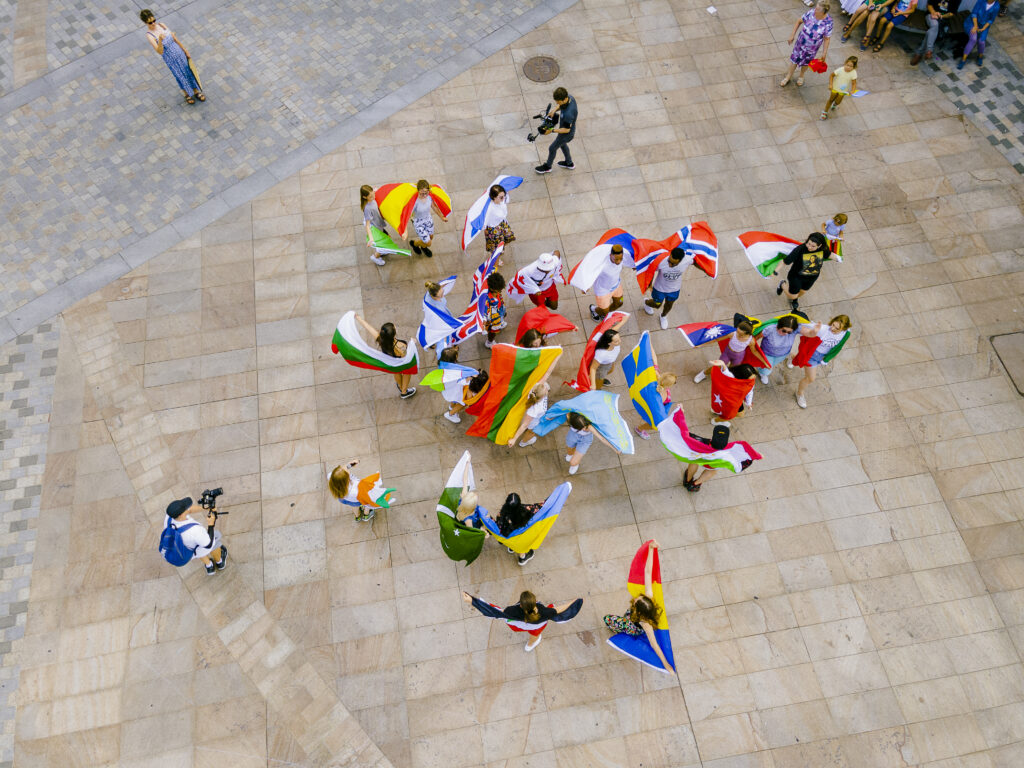 Zdjęcie z góry (z drona). W centrum miasta grupa młodych ludzi tańczy z wielobarwnymi flagami państw, z których pochodzą. 